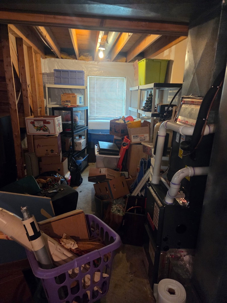 Tight basement mechanical-room walkway crowded with boxes, bins, and stored household items before a cleanout on Buckskin Lake Drive near Glenelg, MD