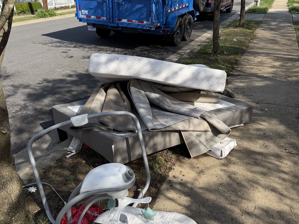 Curbside pile of a mattress, box spring, rug and baby gear with the MD Junk Pros truck before a tenant turnover on Laurel Avenue in Laurel, MD
