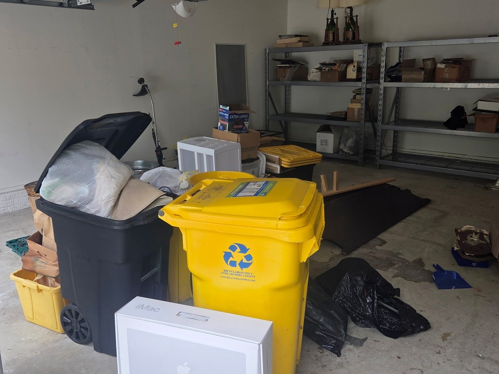 Two-car garage cluttered with full trash and recycling bins, boxes, and old electronics before a cleanout in Millersville, MD
