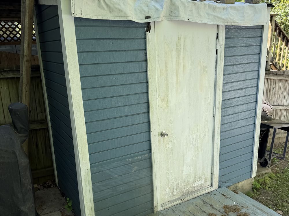Failing resin shed with a tarped roof wedged under deck stairs before removal in Pasadena, MD